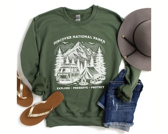 May include: A green sweatshirt with a white graphic design of a vintage camper van, mountains, trees, and a tent. The text on the sweatshirt reads "Discover National Parks Explore - Preserve - Protect".