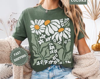 May include: Green t-shirt featuring a floral design with large white daisies with orange centers and smaller white flowers. The shirt has the words "Comfort Colors" printed on it.