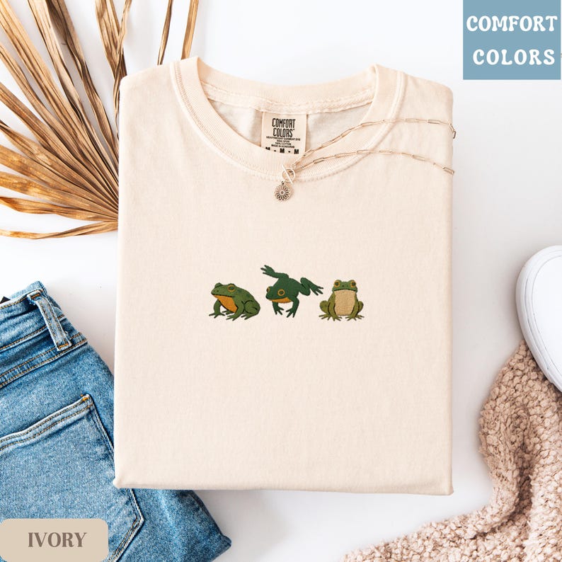 May include: Ivory-colored t-shirt with an embroidered design of three green frogs. The shirt is from Comfort Colors and features a crew neck. A gold necklace with a small pendant is draped on the shirt. The shirt is folded on a surface with blue jeans and a beige sweater.