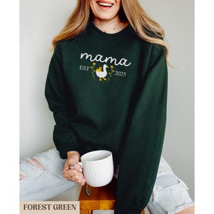 May include: Forest green sweatshirt with the text "mama est 2025" and a white goose with yellow flowers.