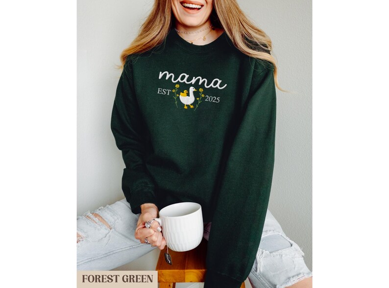 May include: Forest green sweatshirt with the text "mama est 2025" and a white goose with yellow flowers.