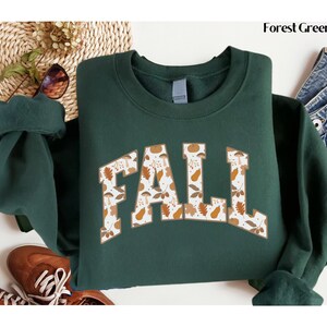 May include: Forest green sweatshirt with the word "FALL" in a collegiate style font. The letters are filled with a pattern of fall leaves, mushrooms, and pears in shades of brown, orange, and white. The sweatshirt is displayed on a white surface.