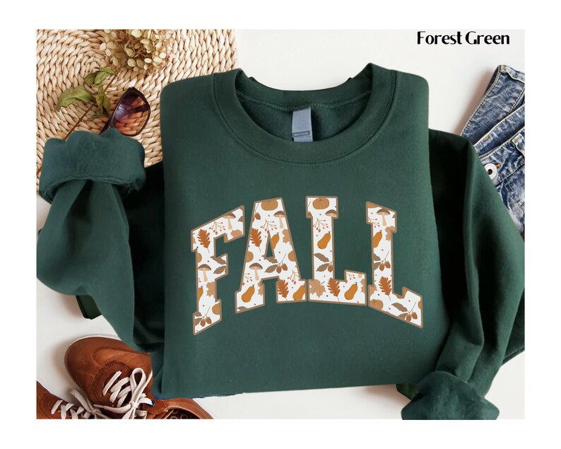 May include: Forest green sweatshirt with the word "FALL" in a collegiate style font. The letters are filled with a pattern of fall leaves, mushrooms, and pears in shades of brown, orange, and white. The sweatshirt is displayed on a white surface.