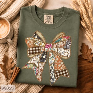 May include: An olive green t-shirt featuring a large, central graphic of a bow constructed from various patchwork fabric patterns, including floral prints, brown and white gingham, and solid colors, all outlined with a gold-toned stitched border.