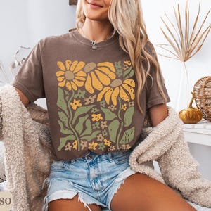 May include: A brown short-sleeve t-shirt featuring a graphic print of yellow sunflowers and green foliage. The design has a distressed texture. The shirt is paired with light blue denim shorts and a textured beige cardigan. The word ESPRESSO is visible.