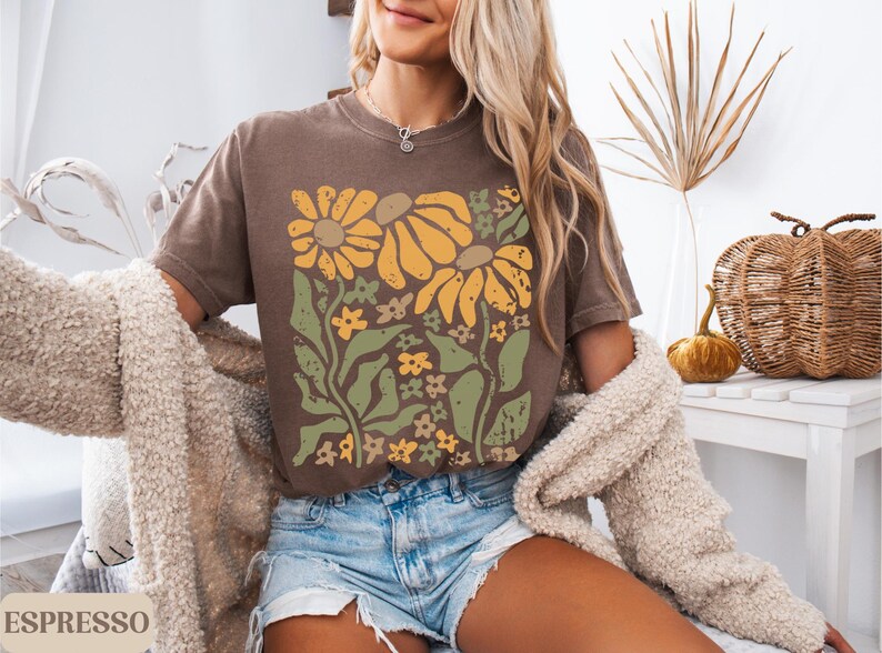 May include: A brown short-sleeve t-shirt featuring a graphic print of yellow sunflowers and green foliage. The design has a distressed texture. The shirt is paired with light blue denim shorts and a textured beige cardigan. The word ESPRESSO is visible.