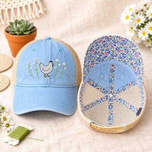 May include: Light blue trucker hat with a chicken and floral embroidery. The hat has a tan mesh back and a floral patterned interior. The hat is a classic baseball cap style.