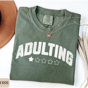 May include: A moss green t-shirt with the word "ADULTING" in white, arched text above five white stars. A gold necklace and a brown hat are also in the image. The shirt is folded on a white surface.