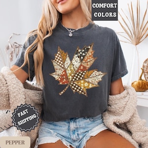 May include: Dark grey t-shirt featuring a patchwork maple leaf design in autumn colors. The leaf design includes floral, plaid, and patterned fabric sections. The words "COMFORT COLORS" are printed at the top. The words "FAST SHIPPING" are printed on a badge.