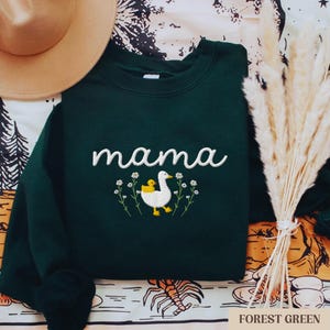 May include: A dark green sweatshirt with the word "mama" embroidered in white and a white duck with a yellow chick embroidered below. The duck is surrounded by white flowers.
