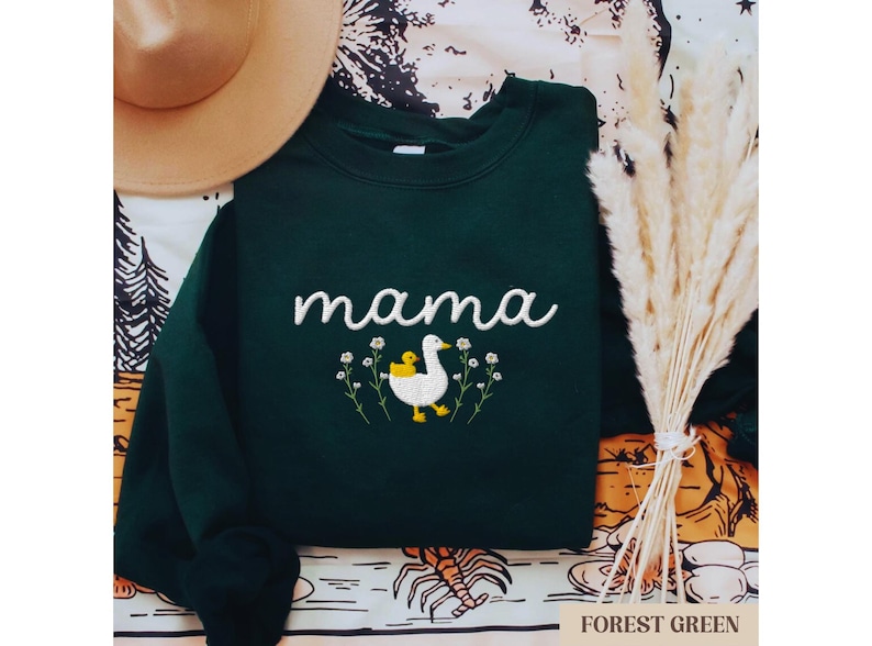 May include: A dark green sweatshirt with the word "mama" embroidered in white and a white duck with a yellow chick embroidered below. The duck is surrounded by white flowers.