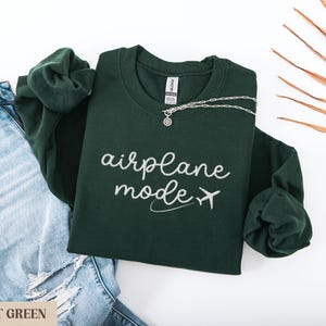 Puede incluir: Sudadera verde bosque con el texto "modo avi&oacute;n" y un peque&ntilde;o avi&oacute;n gr&aacute;fico en blanco.