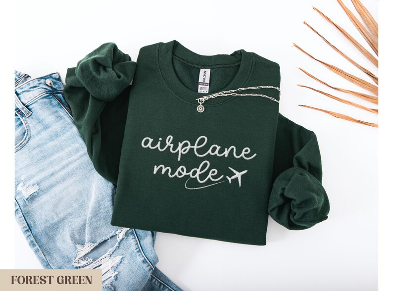 Puede incluir: Sudadera verde bosque con el texto "modo avi&oacute;n" y un peque&ntilde;o avi&oacute;n gr&aacute;fico en blanco.