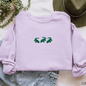 May include: A light purple sweatshirt with three green alligator designs embroidered across the chest. The sweatshirt is folded, with the sleeves bunched up. The word "ORCHID" is printed in the bottom right corner.