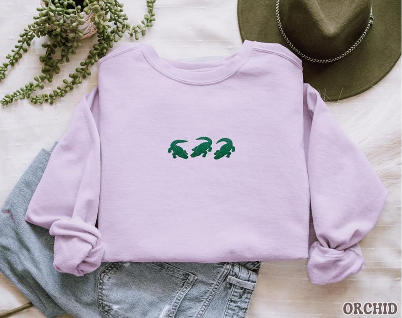 May include: A light purple sweatshirt with three green alligator designs embroidered across the chest. The sweatshirt is folded, with the sleeves bunched up. The word "ORCHID" is printed in the bottom right corner.