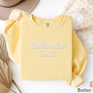 Op de afbeelding: Een lichtgele sweatshirt met de geborduurde tekst "Saltwater Soul" in wit script. De top met lange mouwen heeft een ronde hals en is gemaakt van een zacht materiaal. Het merklabel "Comfort Colors" is zichtbaar op de kraag.