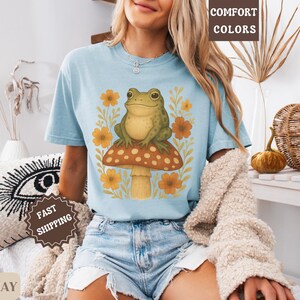 May include: A light blue t-shirt featuring a graphic of a green frog sitting on a mushroom with yellow flowers. The text "COMFORT COLORS" is visible in the top right corner of the image.