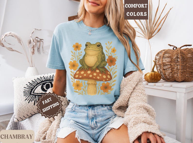 May include: A light blue t-shirt featuring a graphic of a green frog sitting on a mushroom with yellow flowers. The text "COMFORT COLORS" is visible in the top right corner of the image.