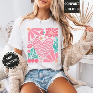May include: White t-shirt featuring a pink heart with a zebra print, surrounded by pink flowers, green leaves, and cherries. The shirt has the words "Comfort Colors" printed on it.