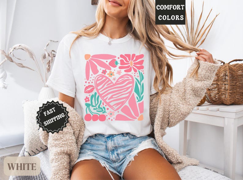 May include: White t-shirt featuring a pink heart with a zebra print, surrounded by pink flowers, green leaves, and cherries. The shirt has the words "Comfort Colors" printed on it.