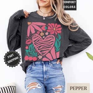 May include: Dark gray long-sleeve sweatshirt with a floral and heart graphic. The design features pink flowers, a zebra-striped heart, cherries, and the word "COMFORT COLORS" in a black box. The sweatshirt is paired with blue jeans.