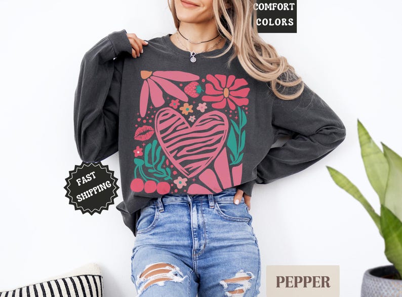May include: Dark gray long-sleeve sweatshirt with a floral and heart graphic. The design features pink flowers, a zebra-striped heart, cherries, and the word "COMFORT COLORS" in a black box. The sweatshirt is paired with blue jeans.