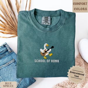 May include: A teal-colored t-shirt with an embroidered design of a duck playing a guitar. The text "SCHOOL OF HONK" is below the duck. The shirt is folded on a white surface next to a pair of blue jeans.