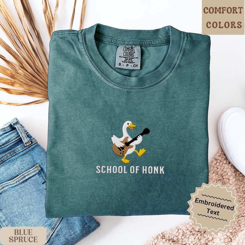 May include: A teal-colored t-shirt with an embroidered design of a duck playing a guitar. The text "SCHOOL OF HONK" is below the duck. The shirt is folded on a white surface next to a pair of blue jeans.