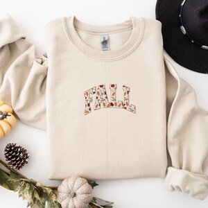 May include: A beige sweatshirt with the word "FALL" printed on the front in a large, bold font. The letters are filled with a pattern of autumn leaves in shades of orange, brown, and red.