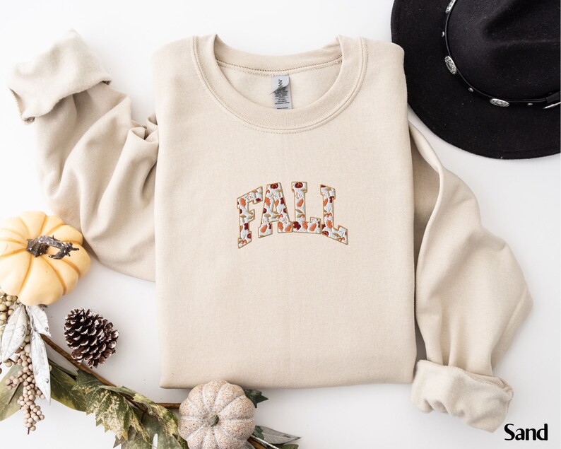 May include: A beige sweatshirt with the word "FALL" printed on the front in a large, bold font. The letters are filled with a pattern of autumn leaves in shades of orange, brown, and red.
