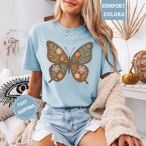 Peut inclure: T-shirt bleu clair orn&eacute; d'un motif papillon marron avec des fleurs et des glands. Le papillon est d&eacute;cor&eacute; d'&eacute;l&eacute;ments orange, roses et verts. La personne porte un short en jean.