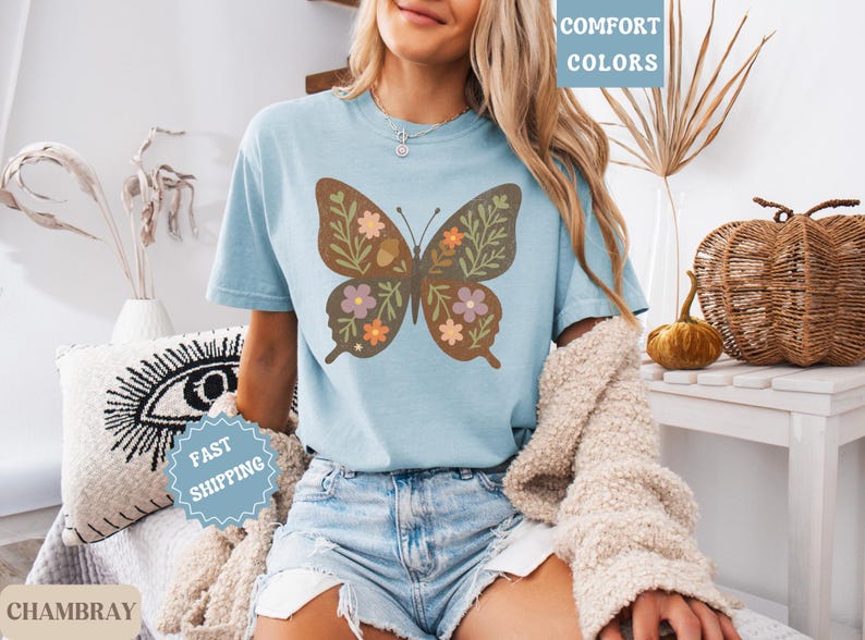 Peut inclure: T-shirt bleu clair orn&eacute; d'un motif papillon marron avec des fleurs et des glands. Le papillon est d&eacute;cor&eacute; d'&eacute;l&eacute;ments orange, roses et verts. La personne porte un short en jean.