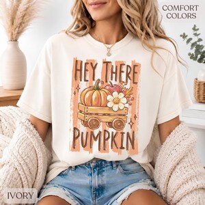 Op de afbeelding: Een ivoorkleurig t-shirt met een grafische print van een houten wagen met een pompoen, een appel en een witte bloem. De tekst "HEY THERE PUMPKIN" is in bruin boven en onder de wagenillustratie gedrukt tegen een achtergrond van oranje penseelstreken.