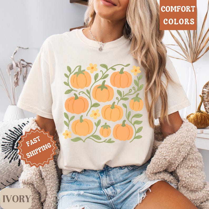 May include: Ivory-colored t-shirt featuring a square pumpkin design with orange pumpkins, green vines, and yellow flowers. The words "Comfort Colors" are printed in a brown box. The words "Fast Shipping" are printed on a brown badge.