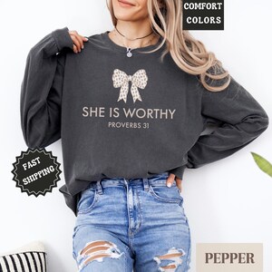 May include: Dark gray long-sleeve sweatshirt with the text "SHE IS WORTHY PROVERBS 31" and a bow graphic. The sweatshirt is a comfort color and is paired with blue jeans. The image also includes a plant and a pillow.