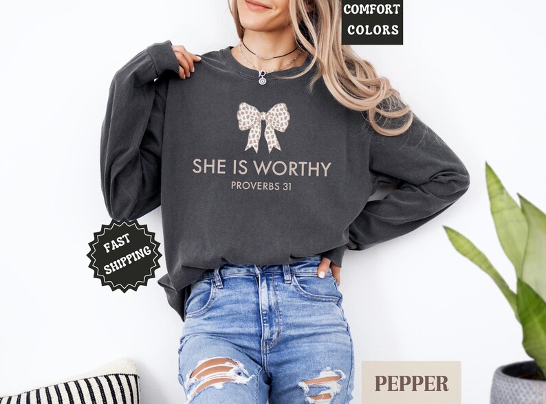 May include: Dark gray long-sleeve sweatshirt with the text "SHE IS WORTHY PROVERBS 31" and a bow graphic. The sweatshirt is a comfort color and is paired with blue jeans. The image also includes a plant and a pillow.