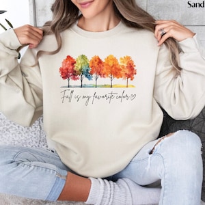 May include: A cream colored sweatshirt with a watercolor design of five trees with fall colored leaves. The text "Fall is my favorite color" is printed below the trees.