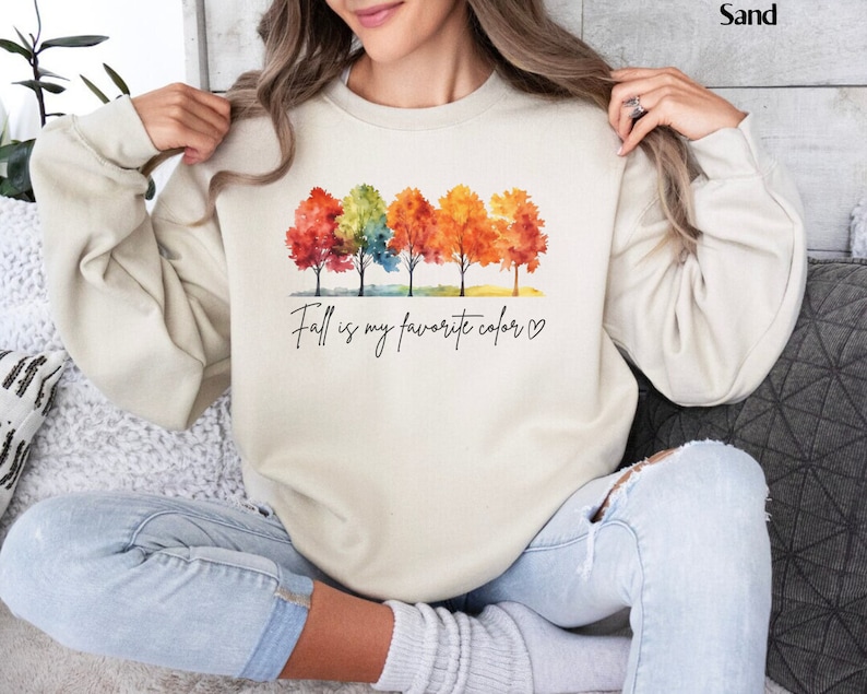 May include: A cream colored sweatshirt with a watercolor design of five trees with fall colored leaves. The text "Fall is my favorite color" is printed below the trees.