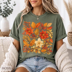May include: A sage green short-sleeve t-shirt featuring a central graphic print of autumn-themed foliage, including orange and red poinsettia flowers, golden yellow leaves, and small orange berries arranged in a square pattern on the chest.