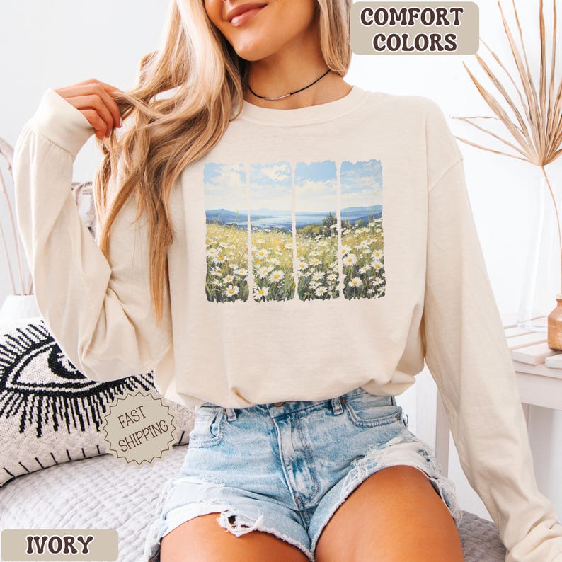 May include: Ivory long-sleeve sweatshirt with a landscape graphic of a field of daisies and a body of water. The words "Comfort Colors" are printed at the top, and "Fast Shipping" is printed on a sticker. The word "Ivory" is printed at the bottom.