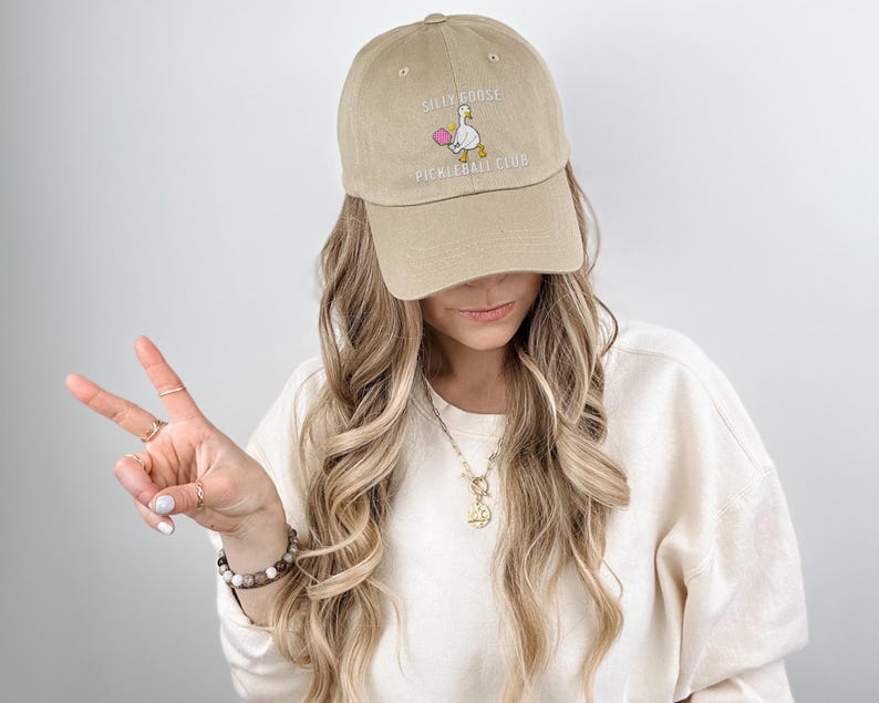 May include: A beige baseball cap with an embroidered white goose holding a pink ball and the text "Silly Goose Pickleball Club".