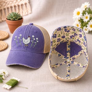 May include: A purple baseball cap with a chicken and floral embroidery. The cap has a mesh back and a floral patterned interior. The cap is displayed next to another cap with the interior visible.