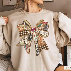 May include: A sand-colored sweatshirt featuring a large, patchwork bow design. The bow incorporates various floral and plaid patterns in shades of orange, brown, and blue. The sweatshirt has long sleeves and a relaxed fit.