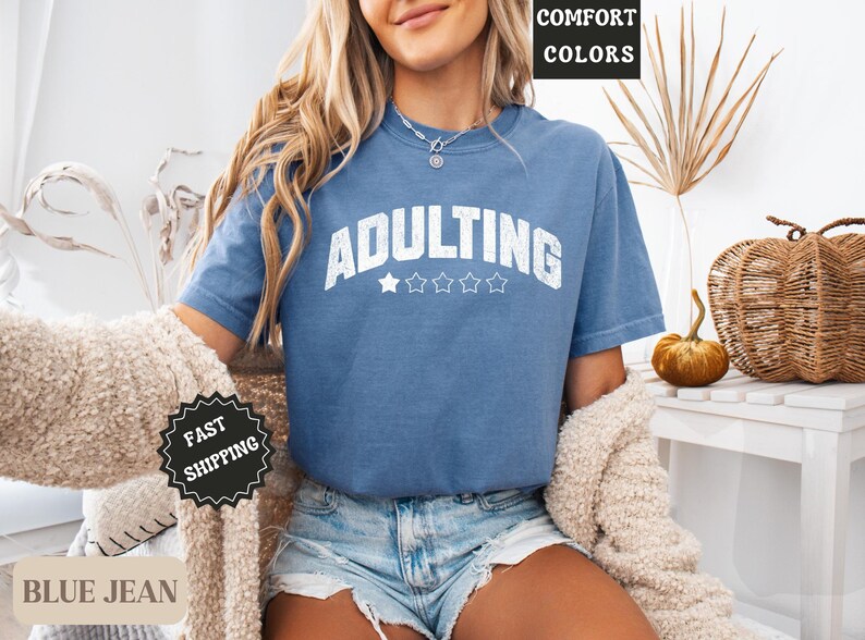 May include: A blue jean-colored t-shirt with the word "ADULTING" in white block letters and five stars underneath. The shirt is part of a comfort colors collection. The model is wearing denim shorts and a cream-colored cardigan.