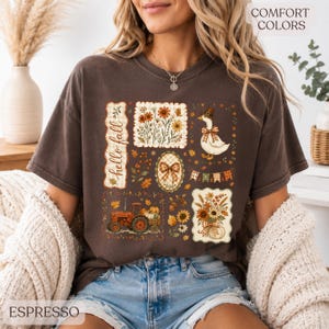 May include: A dark brown short-sleeve t-shirt featuring a graphic print with the text "hello fall". The design includes a tractor, a goose wearing a hat, floral arrangements, pumpkins, and autumn leaves in shades of orange, yellow, and brown.
