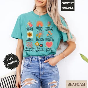 Pu&ograve; includere: Una maglietta blu turchese con un design floreale e affermazioni positive stampate sul davanti. Il testo include "be kind always", "choose joy", "keep on blooming", "you matter", "focus on the good", "growth is a process", "the world needs you", "life is beautiful", and "you are loved".