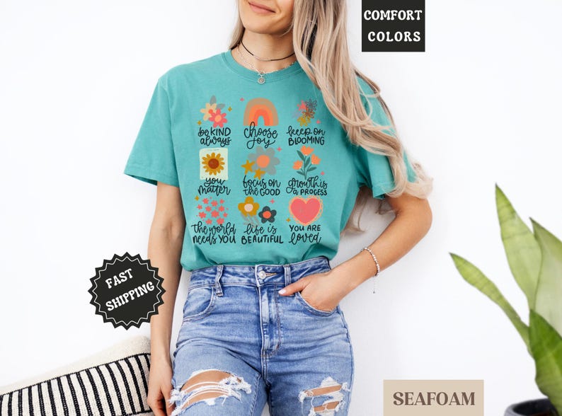 Pu&ograve; includere: Una maglietta blu turchese con un design floreale e affermazioni positive stampate sul davanti. Il testo include "be kind always", "choose joy", "keep on blooming", "you matter", "focus on the good", "growth is a process", "the world needs you", "life is beautiful", and "you are loved".