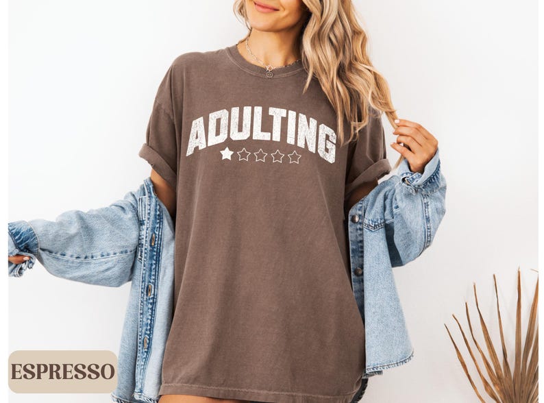 May include: Espresso-colored t-shirt with the word "ADULTING" in distressed white lettering and five stars. The shirt is worn with a light blue denim jacket.