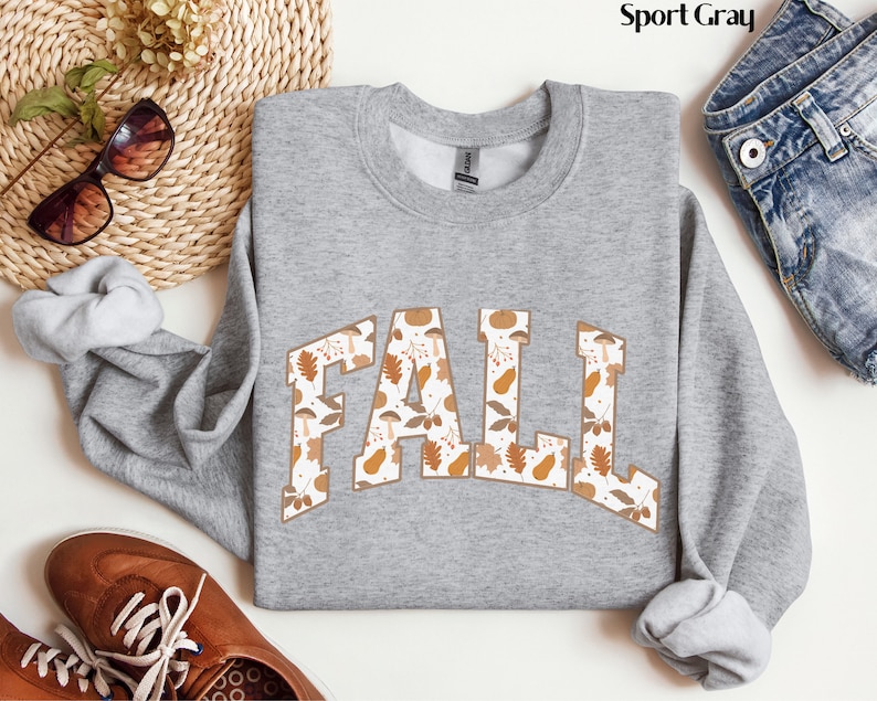 May include: A light gray sweatshirt with the word "FALL" in a brown, fall-themed pattern. The letters are filled with images of leaves, mushrooms, and pumpkins. The sweatshirt is displayed with brown shoes, sunglasses, and denim jeans.