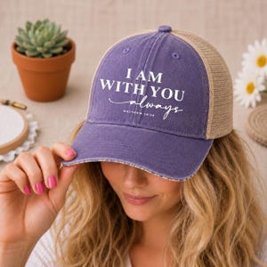 May include: A purple and tan baseball cap with the text "I AM WITH YOU always" in white. The cap has a mesh back and a curved brim. The text "Matthew 28:20" is below the main text.